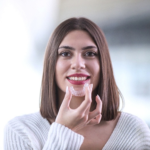 A young woman with brown hair prepares to insert an Invisalign aligner into her mouth to shift her teeth